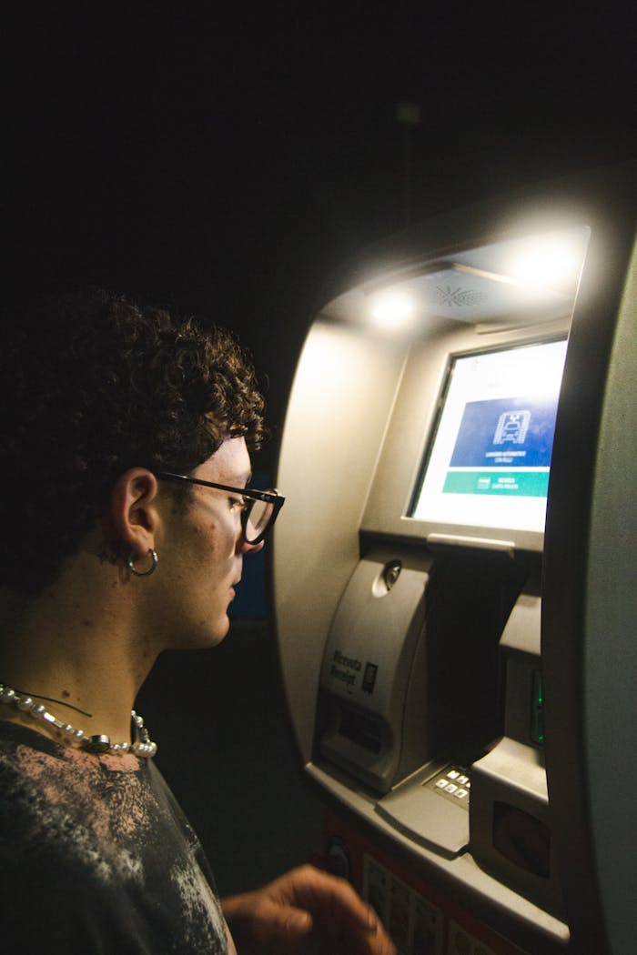 A man operates an ATM machine at night, outdoors under bright lighting.