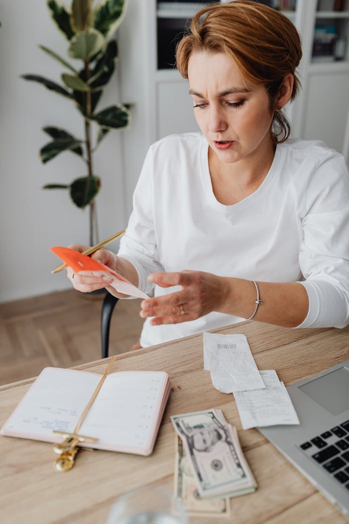 Woman calculating expenses with calculator and notebook at home office, surrounded by receipts and money.